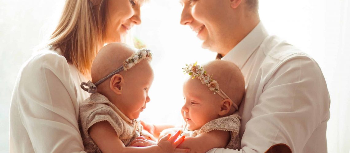 Happy mom and dad pose with funny twins on their arms before a bright window